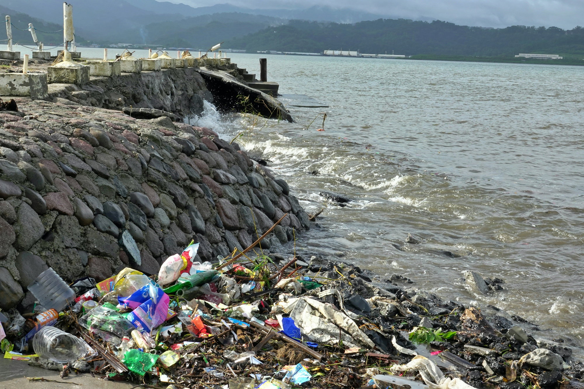 Trash litters the shoreline next to a stone wall.